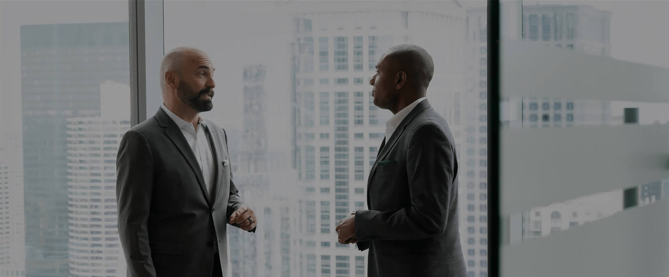 Two men in suits stand talking by a large window overlooking a city skyline.